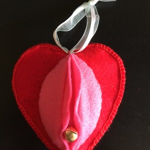 May include: A red felt heart-shaped ornament with a pink felt interior and a gold bell. The ornament has a white ribbon for hanging.