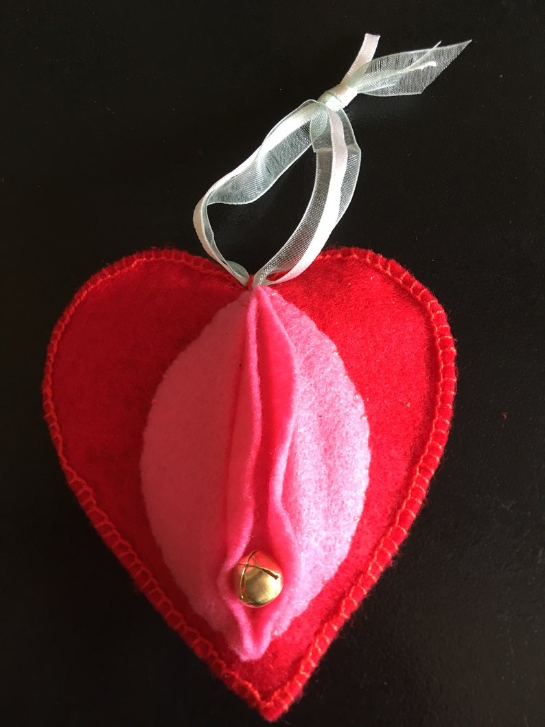 May include: A red felt heart-shaped ornament with a pink felt interior and a gold bell. The ornament has a white ribbon for hanging.