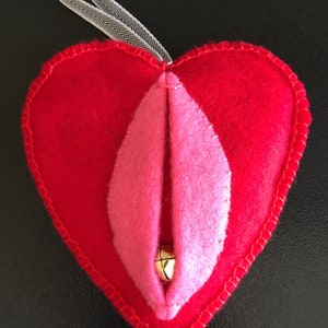 May include: A red felt heart-shaped ornament with a pink felt interior and a gold bell in the center. The ornament has a white ribbon attached to the top.