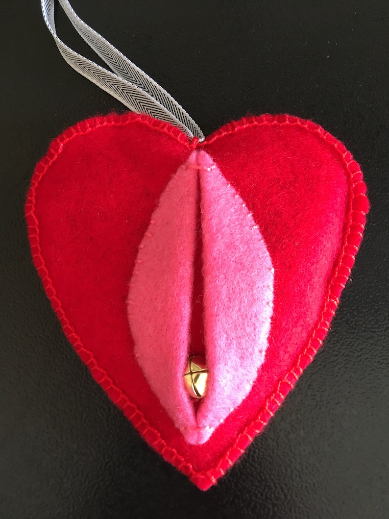 May include: A red felt heart-shaped ornament with a pink felt interior and a gold bell in the center. The ornament has a white ribbon attached to the top.