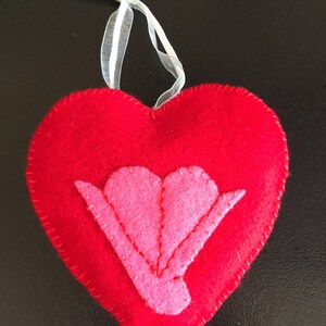May include: A red felt heart with a pink felt flower in the center. The heart has a white ribbon attached to the top.