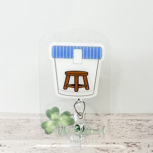 Può includere: Un portabadge bianco e blu con un'immagine a cartoni animati di uno sgabello all'interno di un contenitore. Il portabadge è attaccato a un supporto in acrilico trasparente con un quadrifoglio e il testo "The Glitteral Shamrock".