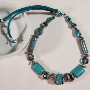 May include: A turquoise and silver necklace with a suede cord. The necklace features turquoise and silver beads, including a large turquoise bead with a silver design.