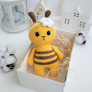 May include: A handmade yellow and brown crocheted bee doll with a white flower headband. The bee is in a white box with straw, and has black eyes and a small "T" on its face. The doll is a cute, whimsical toy.