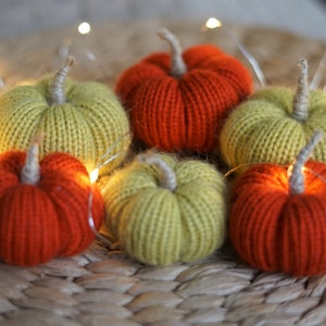 May include: Six knitted pumpkins, three green and three orange, arranged on a woven surface with string lights.