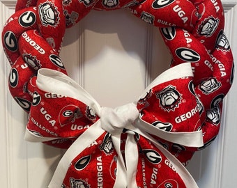 May include: A red and white braided wreath with a white bow. The wreath is decorated with the University of Georgia Bulldogs logo and the words "UGA" and "Georgia Bulldogs".
