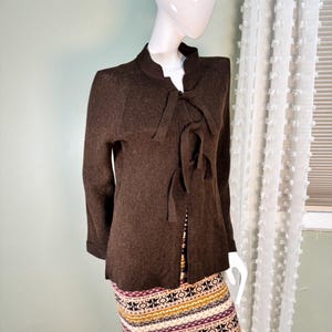 May include: A brown long-sleeve sweater with a bow detail at the neckline. The sweater has a high collar and is displayed on a mannequin. The sleeves are cuffed at the wrists. The mannequin is wearing a patterned skirt.
