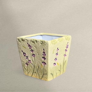 Puede incluir: Maceta cuadrada de cerámica con acabado amarillo claro y diseño floral de lavanda. La maceta presenta ramitas de lavanda en relieve en morado y verde, con una superficie texturizada. El interior es blanco.