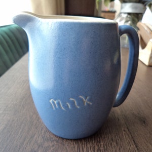 Puede incluir: Una jarra de leche de cerámica azul con una manija blanca y la palabra "Leche" inscrita en la parte delantera.