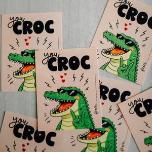 May include: A collection of greeting cards with a cartoon alligator wearing sunglasses. The cards feature the text "you CROC" in black, with lightning bolts and hearts. The alligator is green with an open mouth.