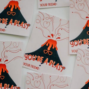 May include: A set of five greeting cards featuring a cartoon volcano with sunglasses and the text "You're a blast" and "Your friend:". The volcano is erupting with orange lava and red hearts.