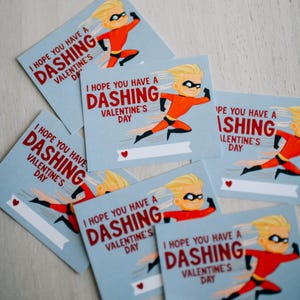 Puede incluir: Tarjetas de San Valentín azul claro con un superhéroe de dibujos animados en rojo y negro. Las tarjetas dicen "I HOPE YOU HAVE A DASHING VALENTINE'S DAY" en texto rojo. Un pequeño corazón rojo en la parte inferior.