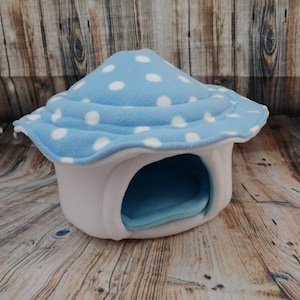 May include: A whimsical pet bed shaped like a mushroom. The mushroom cap is light blue with white polka dots, and the base is white. The interior is light blue, providing a cozy space for small pets. The bed is made of soft fleece.