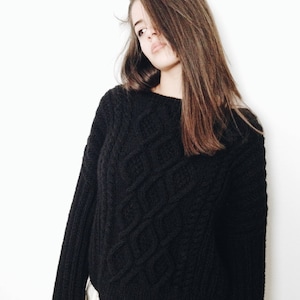 Puede incluir: Un suéter negro de punto con un patrón de diamantes. El suéter tiene mangas largas y cuello redondo. La prenda está hecha de un hilo grueso y texturizado. El suéter se combina con una falda blanca. El fondo es una pared blanca lisa.