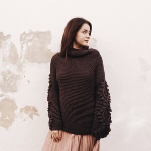 Puede incluir: Un suéter de cuello alto marrón con tejido texturizado y grandes detalles redondos texturizados en las mangas.