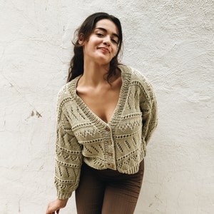 May include: A woman wearing a light green crocheted cardigan with a v-neck and button closure. The cardigan has a delicate openwork pattern. She is wearing brown trousers.