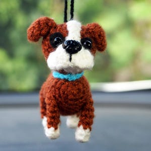 Puede incluir: Adorno de perro tejido a ganchillo, marrón y blanco, con un collar azul. El perro tiene ojos negros y nariz negra. Cuelga de un hilo negro, adecuado para decorar un coche o una habitación. El fondo está borroso.