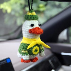 Op de afbeelding: Een gehaakt eend-ornament dat aan een zwart koord hangt. De eend heeft een witte kop, een oranje snavel en pootjes, en draagt een groene muts met een gele band, een groene trui met een gele letter O en een felgele sjaal om de nek geknoopt.