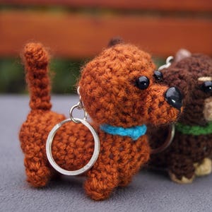 May include: A collection of crocheted dachshund keyrings in shades of brown, dark brown, and beige. Each keyring has a black nose, black eyes, and a collar. The keyrings are attached to a silver metal ring.