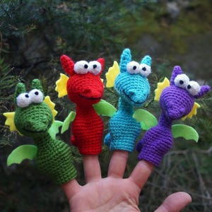 May include: Four colorful crocheted finger puppets shaped like dragons. The dragons are green, red, blue, and purple, each with white eyes, yellow accents, and green wings. The puppets are displayed on fingers against a blurred green background.