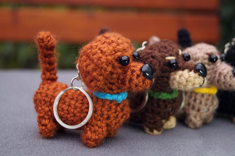May include: A collection of crocheted dachshund keyrings in shades of brown, dark brown, and beige. Each keyring has a black nose, black eyes, and a collar. The keyrings are attached to a silver metal ring.