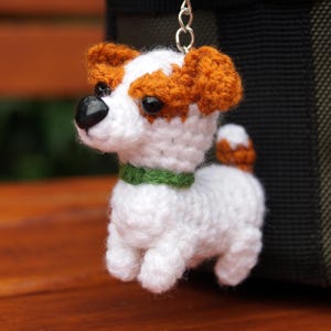 Op de afbeelding: Een handgemaakte gehaakte hondenbedel in wit, bruin en zwart. De hond heeft een groene halsband en een zwarte neus. De bedel is opgehangen aan een zilverkleurige metalen lus. De hond is een Jack Russell Terrier.