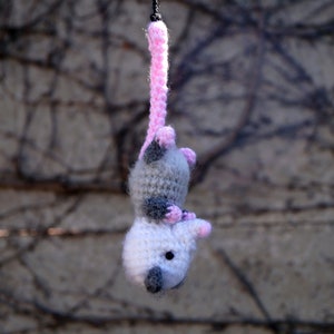 May include: A crocheted opossum toy hanging from a pink string. The opossum is gray and white with pink accents.