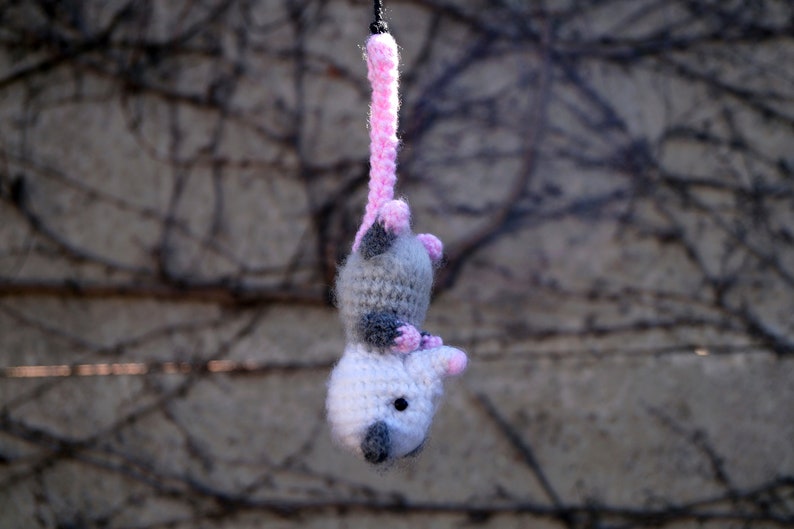 May include: A crocheted opossum toy hanging from a pink string. The opossum is gray and white with pink accents.