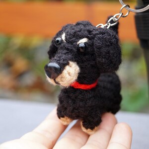 May include: A crocheted black and tan dachshund keyring with a red collar. The small dog has a black nose and button eyes. The keyring is attached to a silver ring.