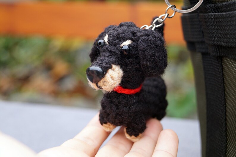 May include: A crocheted black and tan dachshund keyring with a red collar. The small dog has a black nose and button eyes. The keyring is attached to a silver ring.