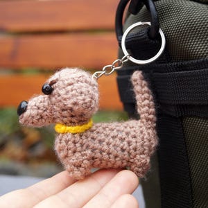May include: A crocheted dachshund dog keyring in brown, with a yellow collar and black features. The dog is attached to a silver keyring. The keyring is held in front of a green bag.