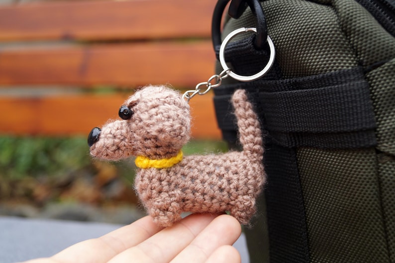 May include: A crocheted dachshund dog keyring in brown, with a yellow collar and black features. The dog is attached to a silver keyring. The keyring is held in front of a green bag.