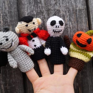 May include: Crocheted Halloween finger puppets including a gray bat, a vampire with a red cape, a skeleton, and an orange pumpkin. The puppets are handmade with detailed features and are displayed on fingers.