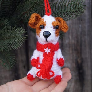 May include: A handmade crochet dog ornament with a red scarf and snowflake detail. The dog has brown and white fur and a black nose. The ornament is hanging from a red string, perfect for holiday decor.