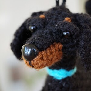 以下が含まれることがあります： 青い首輪をした黒と茶色の毛糸で編まれた犬のぬいぐるみ。おもちゃは黒い目と茶色の鼻をしています。