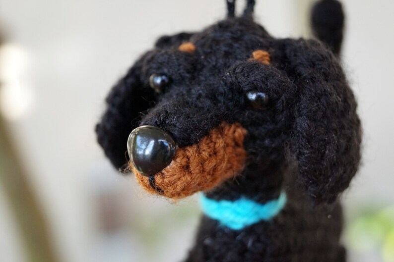 以下が含まれることがあります： 青い首輪をした黒と茶色の毛糸で編まれた犬のぬいぐるみ。おもちゃは黒い目と茶色の鼻をしています。
