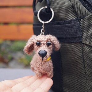 May include: A crocheted dog keyring in shades of brown, with a black nose and black eyes. The dog has a yellow collar and is attached to a silver keyring. The keyring is hanging next to a green bag.