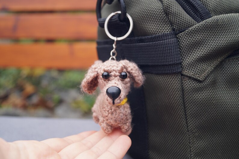 May include: A crocheted dog keyring in shades of brown, with a black nose and black eyes. The dog has a yellow collar and is attached to a silver keyring. The keyring is hanging next to a green bag.