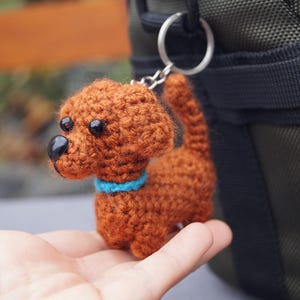 May include: A crocheted dog keyring in shades of brown, with a blue collar and black eyes. The handmade dog is attached to a silver keyring. The background is blurred, with a glimpse of a green bag.