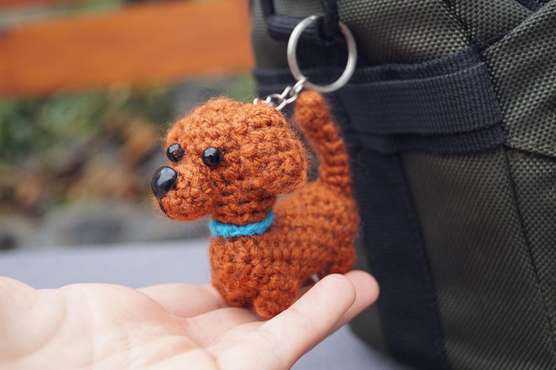 May include: A crocheted dog keyring in shades of brown, with a blue collar and black eyes. The handmade dog is attached to a silver keyring. The background is blurred, with a glimpse of a green bag.