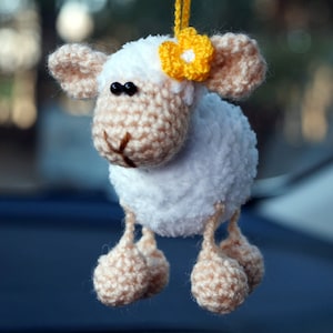 Puede incluir: Un cordero de crochet blanco con una flor amarilla en la cabeza. El cordero tiene ojos negros y una nariz rosa. Está colgado de una cuerda.