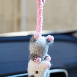 May include: A crocheted opossum hanging from a rearview mirror. The opossum is gray and white with pink accents. It is a cute and whimsical car charm.