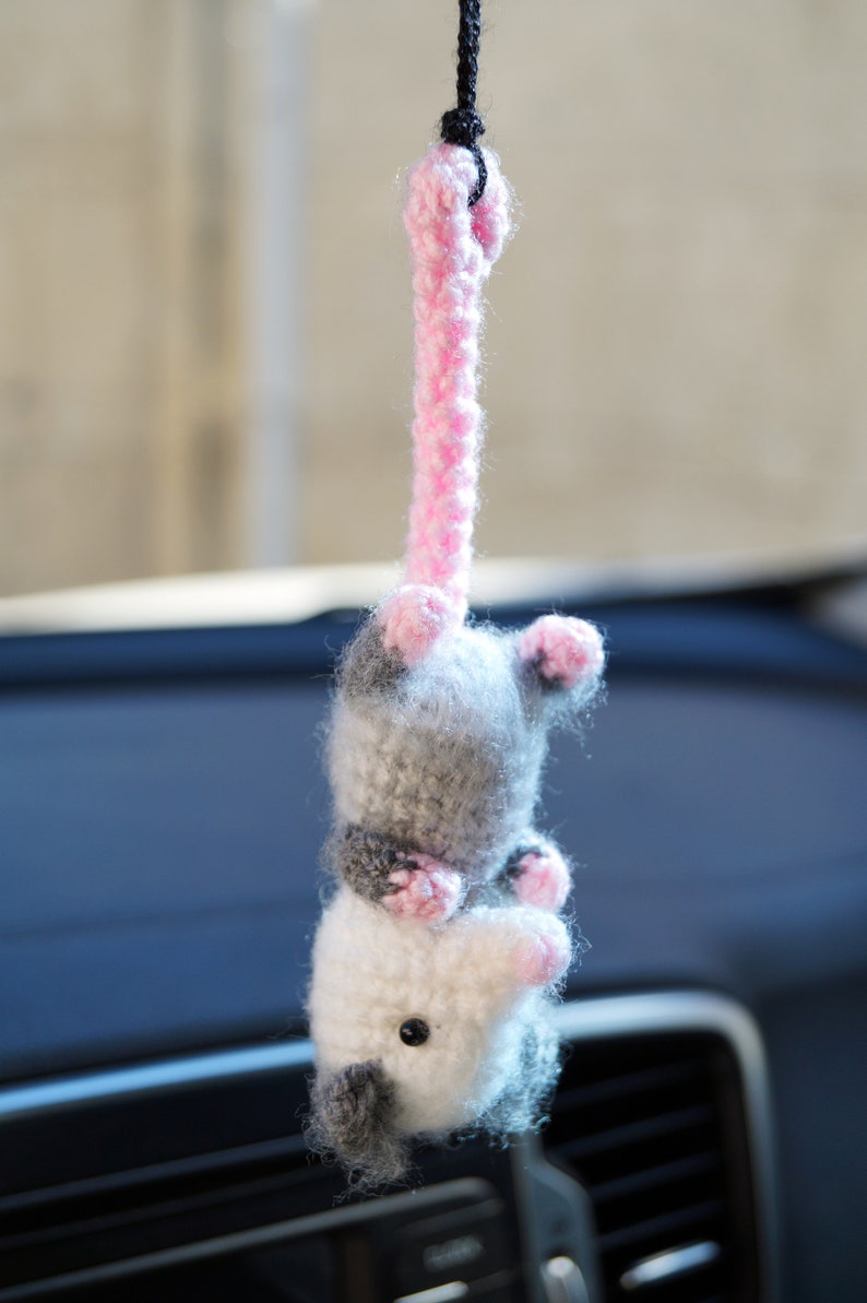 May include: A crocheted opossum hanging from a rearview mirror. The opossum is gray and white with pink accents. It is a cute and whimsical car charm.