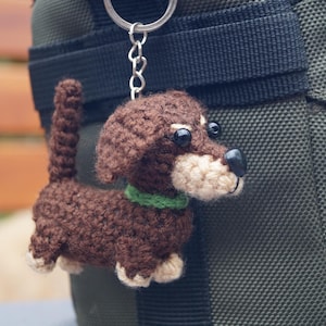 May include: A crocheted dachshund dog keyring. The dog is brown with a cream-coloured snout, paws, and ears. It has black eyes and a green collar. The keyring is attached to a silver chain.