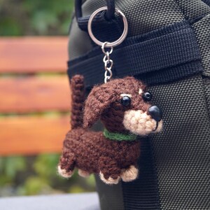 May include: A crocheted dachshund dog keyring. The dog is brown with a beige muzzle and paws, and a green collar. It hangs from a silver-coloured keyring attached to a dark green bag.