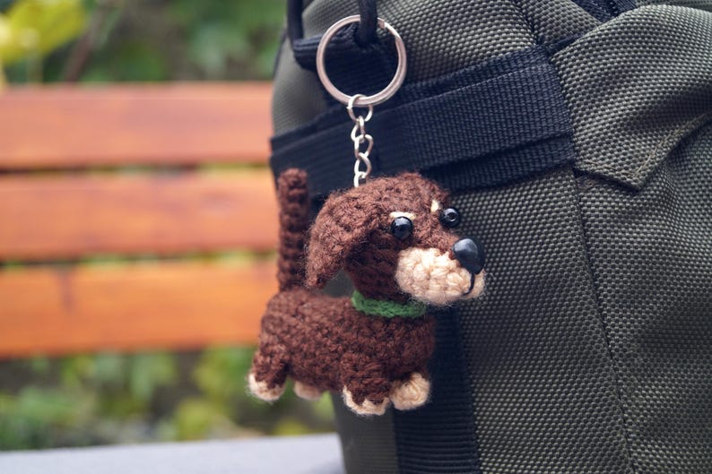 May include: A crocheted dachshund dog keyring. The dog is brown with a beige muzzle and paws, and a green collar. It hangs from a silver-coloured keyring attached to a dark green bag.
