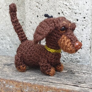 May include: A brown crocheted dachshund dog toy with a yellow collar. The dog has a brown nose and black eyes.