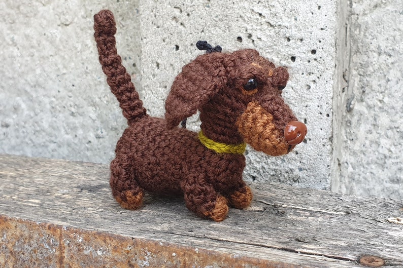 May include: A brown crocheted dachshund dog toy with a yellow collar. The dog has a brown nose and black eyes.