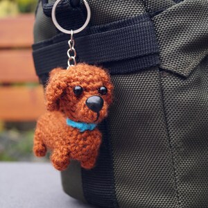 May include: A handmade, brown crochet dog keyring with a blue collar, attached to a silver ring. The dog has black eyes and a black nose. The keyring hangs next to a dark green bag.