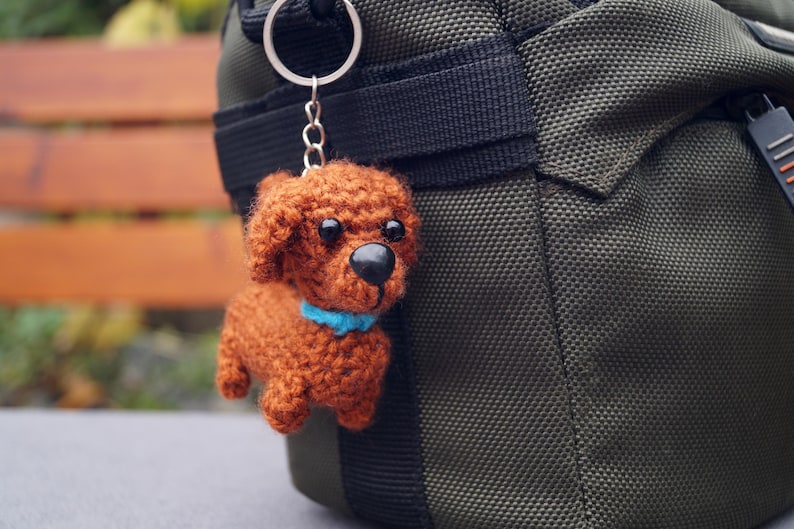 May include: A handmade, brown crochet dog keyring with a blue collar, attached to a silver ring. The dog has black eyes and a black nose. The keyring hangs next to a dark green bag.